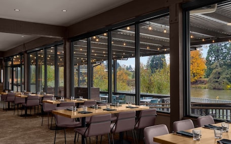 A restaurant with large windows offering views of a river and autumn trees. Tables are set with chairs and glasses.