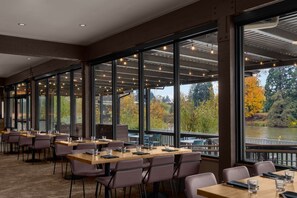 A restaurant with large windows offering views of a river and autumn trees. Tables are set with chairs and glasses.