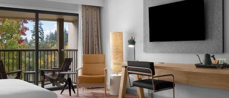 A hotel room with a bed, a desk, a chair, a TV, and a balcony with a view of trees.