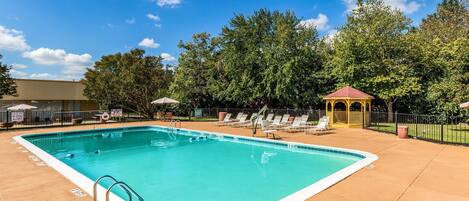 Una piscina rectangular con borde blanco, rodeada por un solárium de hormigón. Se observan sillones de descanso y un pabellón al fondo.