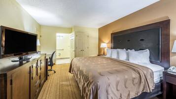 A hotel room with a large bed, a television on a wooden cabinet, a desk with a lamp, and a door leading to another room.