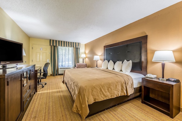 A hotel room with a large bed, a desk with a flat-screen TV, a chair, a nightstand with a lamp, and a window with curtains.
