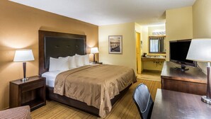 A hotel room with a large bed, two bedside tables with lamps, a desk with a TV, and a small kitchenette.