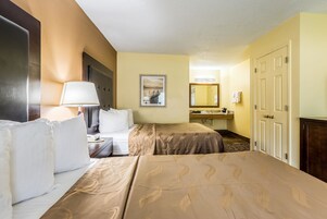 Habitación de hotel con dos camas, una mesita de noche con lámpara, un baño con espejo y una puerta.