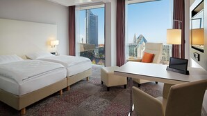 Habitación de hotel con dos camas, un escritorio con una computadora portátil, una silla y ventanas amplias con vista a la ciudad.