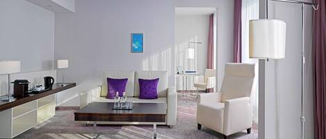 A modern hotel room with a sofa, armchair, coffee table, and a desk with a lamp.