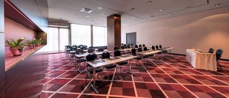 Un salón de conferencias con varias mesas y sillas, piso cubierto de alfombra roja y amplias ventanas con persianas.