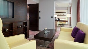 Una habitación de hotel moderna con una televisión de pantalla plana, un mueble de madera oscura con estantes, un sofá color crema con cojines morados y una pequeña mesa de centro.