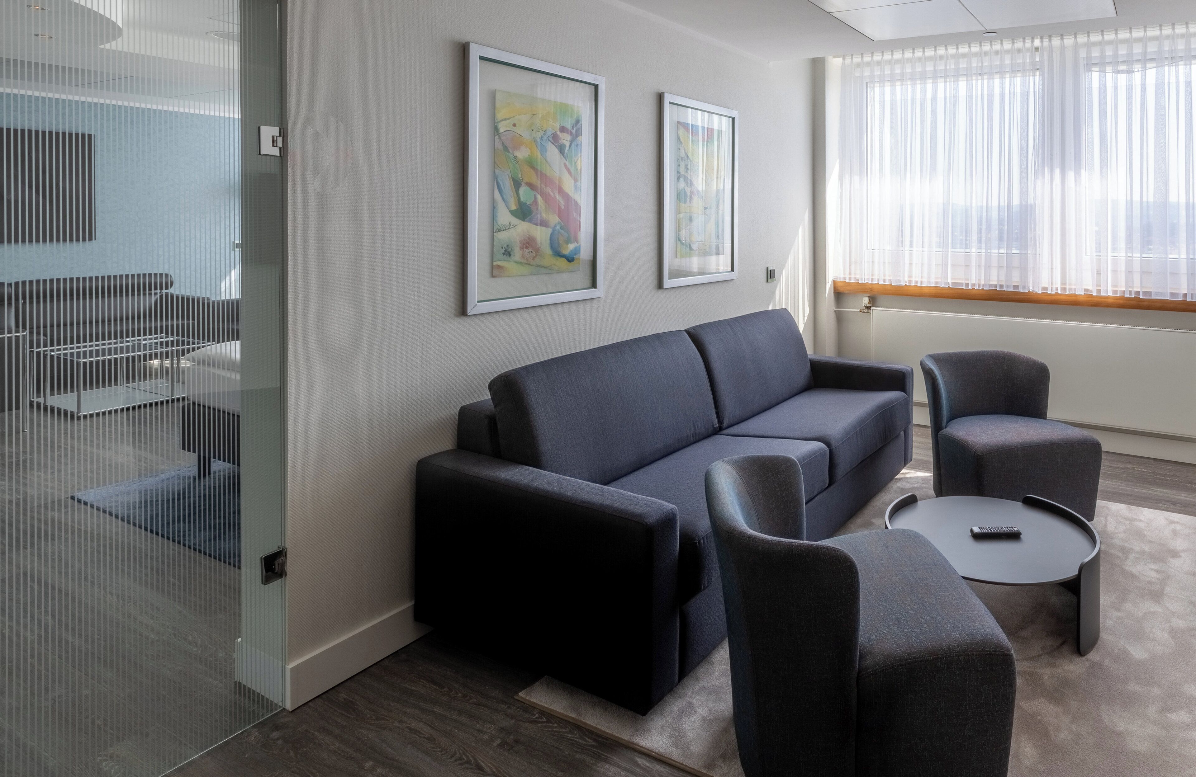 A modern living room with a dark blue sofa, two armchairs, and a small coffee table. There are two framed abstract paintings on the wall, a large window with sheer curtains, and a glass partition with a metal railing.