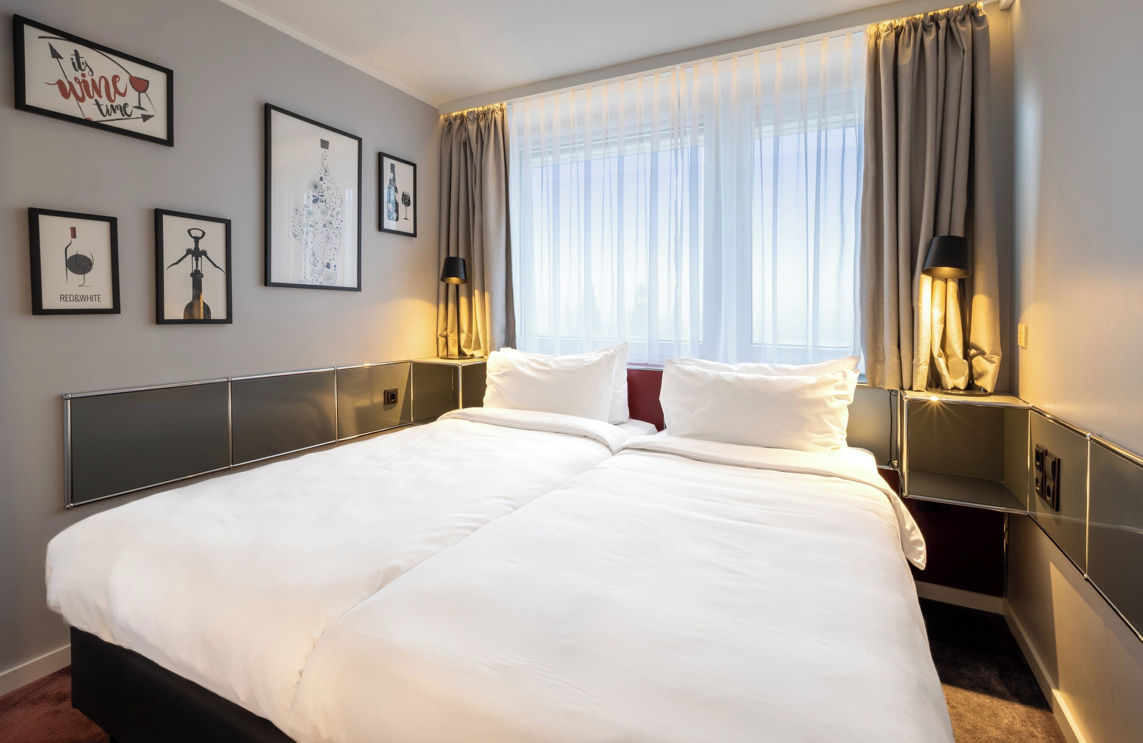A hotel room with a large bed, two bedside tables with lamps, a window with sheer curtains, and several framed pictures on the wall.