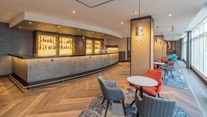 A modern bar area with a curved counter, seating with patterned cushions, and a large window providing natural light.