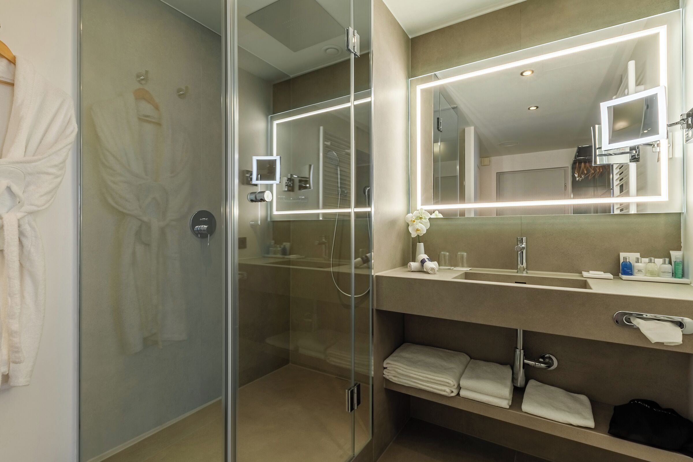 A modern bathroom with a glass-enclosed shower, a large mirror with integrated lighting, and a vanity with a sink and towel storage.