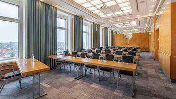 Ein Konferenzraum mit mehreren Tischen und Stühlen, großen Fenstern mit Vorhängen und Blick auf die Stadt.