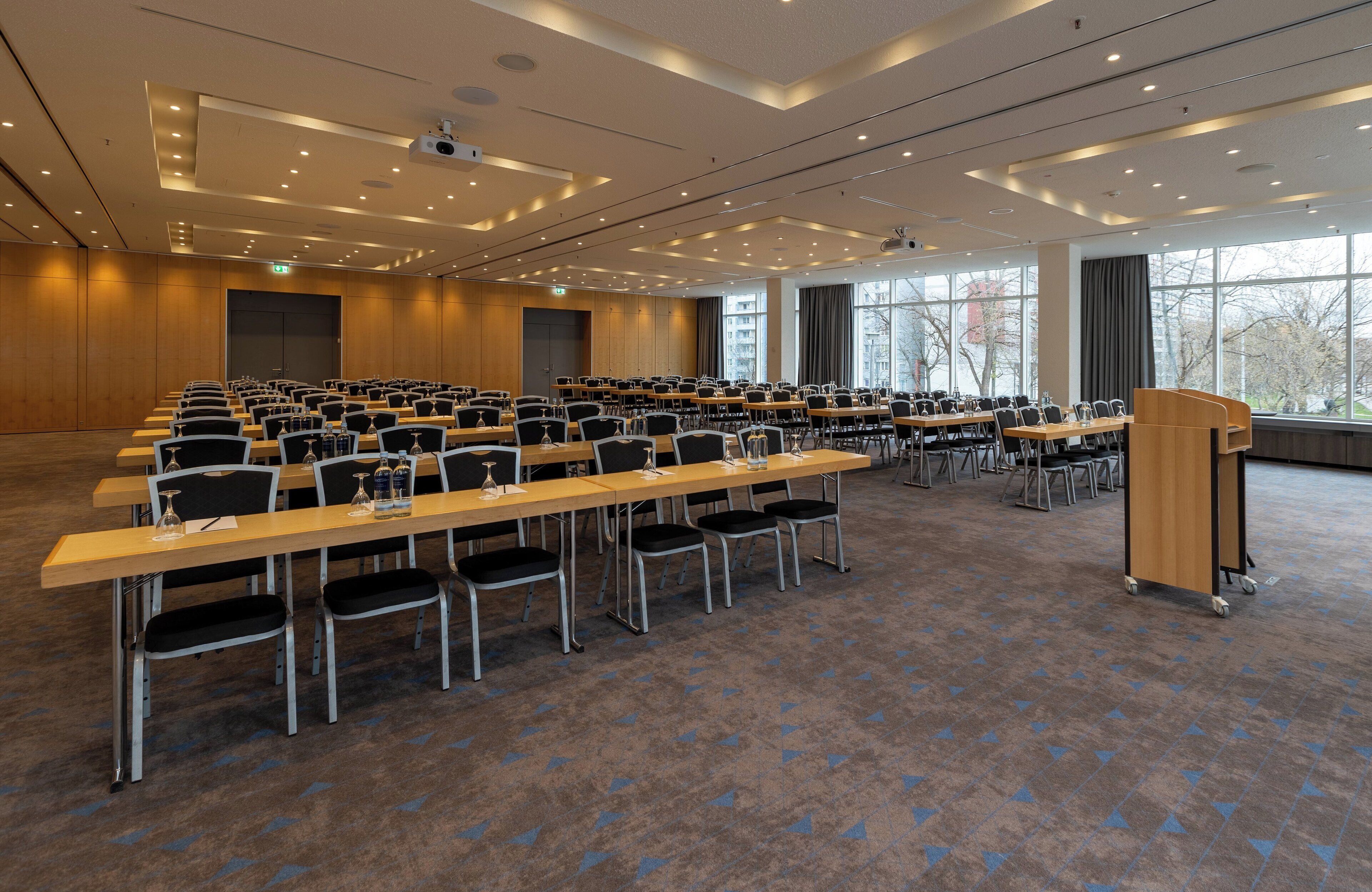 A conference room with rows of tables and chairs, a podium, and large windows with a view of buildings.