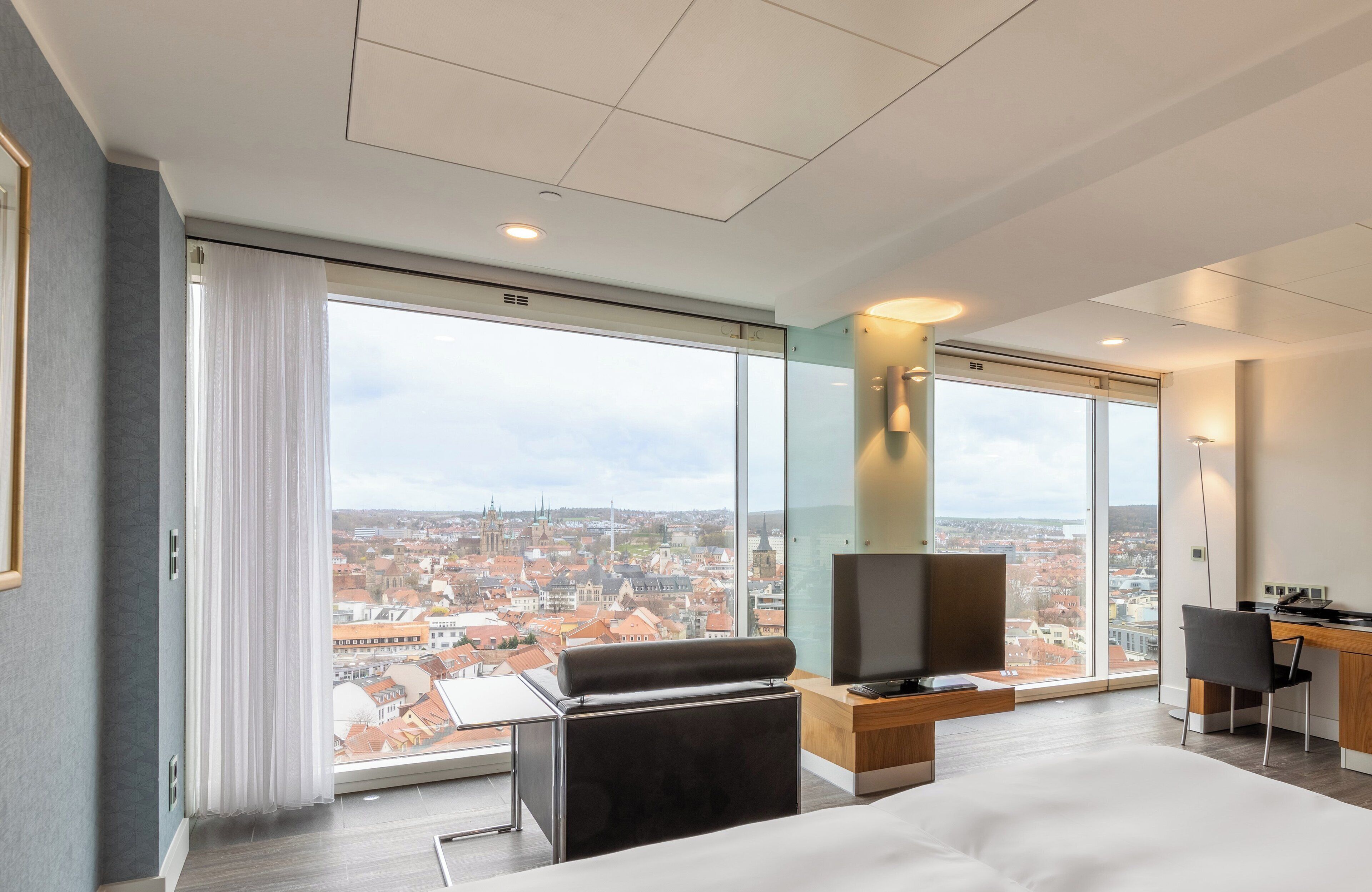 A modern hotel room with a large window offering a city view, a bed, a desk with a chair, and a television.