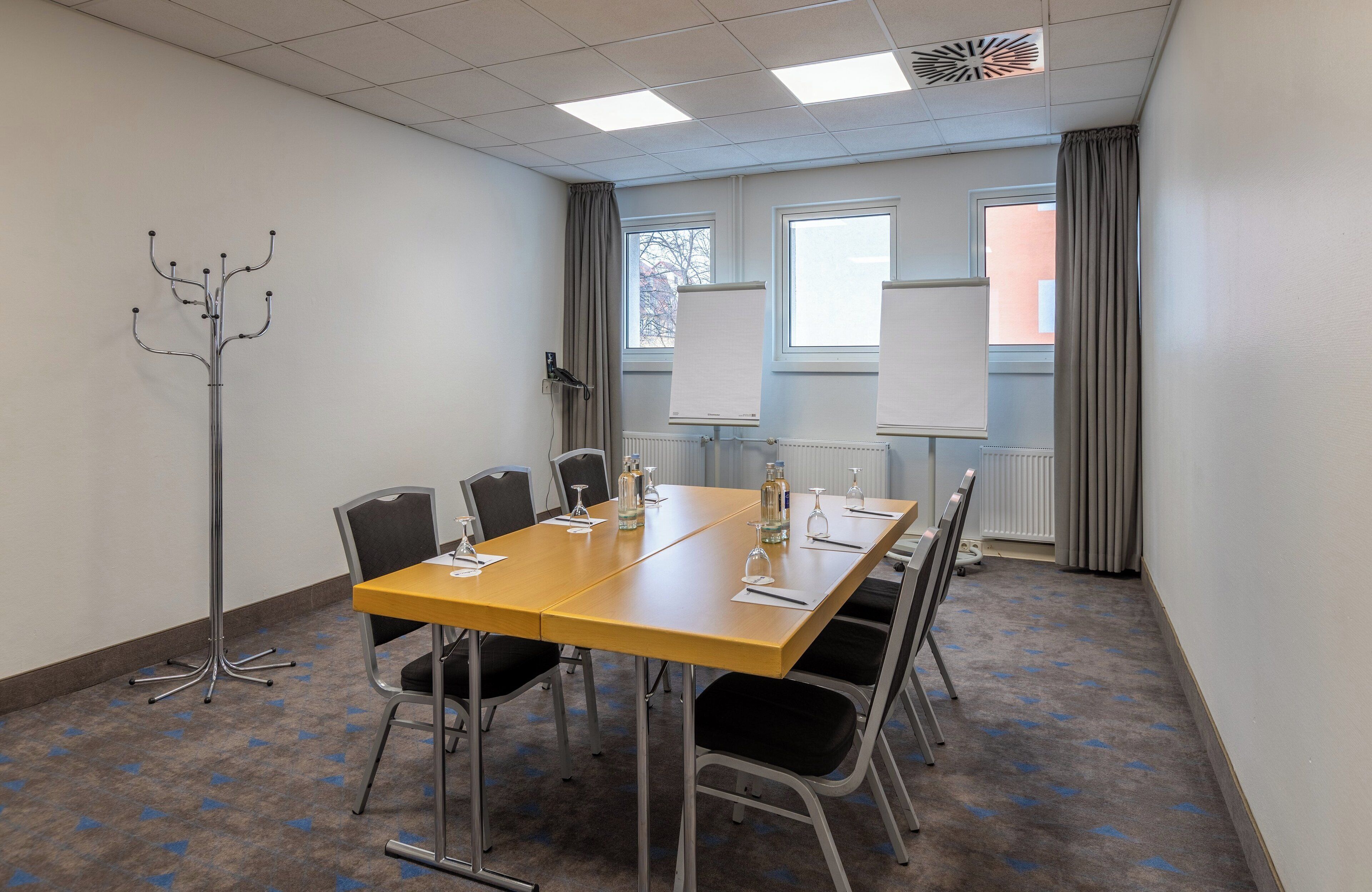 A conference room with a long table, chairs, a coat rack, and windows with curtains.