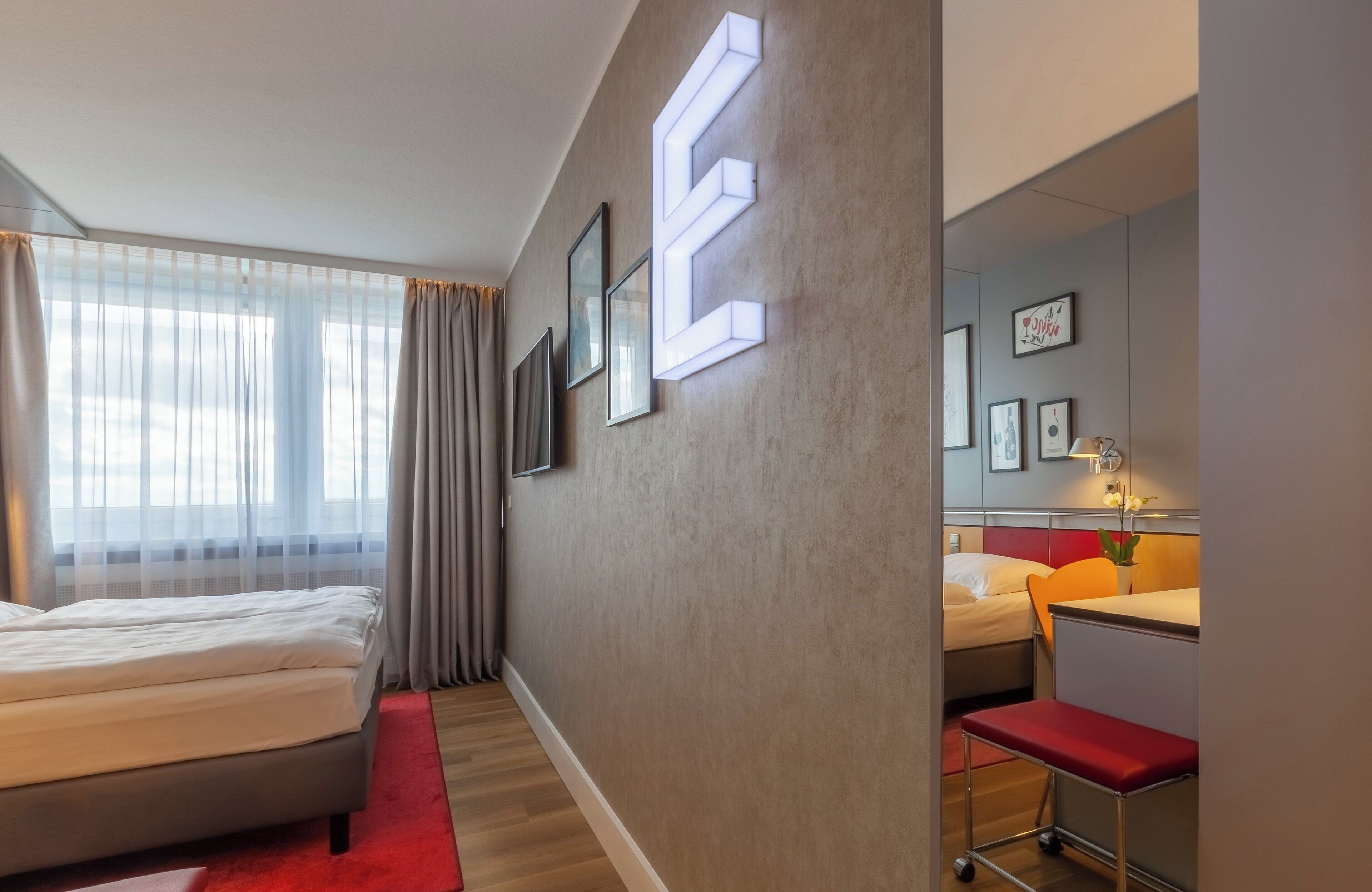 A modern hotel room with a large bed, a desk, and a chair. There are framed pictures on the wall and a large illuminated 'E' on the wall.