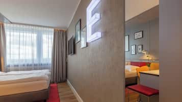 A modern hotel room with a large bed, a desk, and a chair. There are framed pictures on the wall and a large illuminated 'E' on the wall.