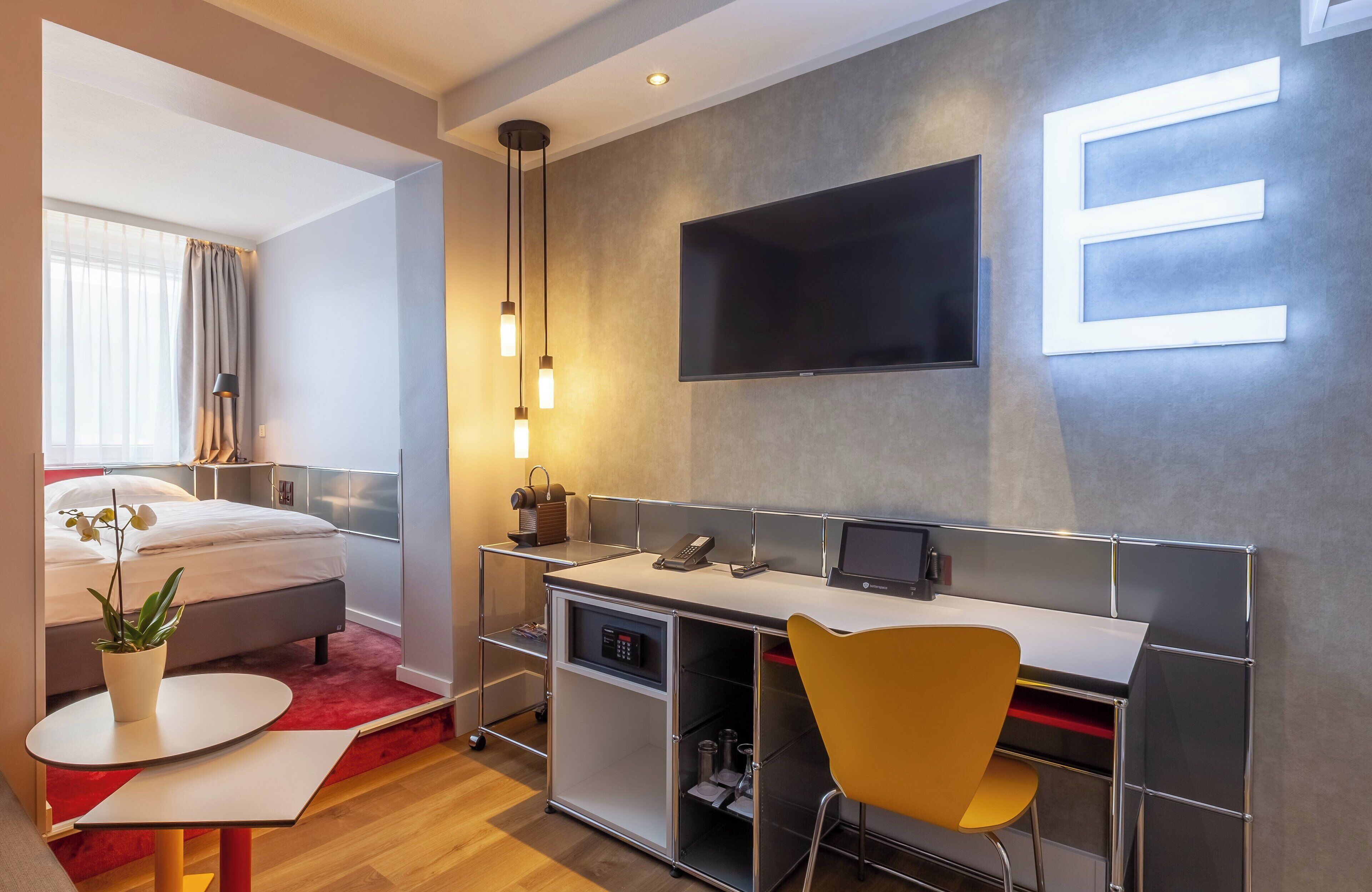 A modern hotel room with a bed, desk, chair, and a TV mounted on the wall.