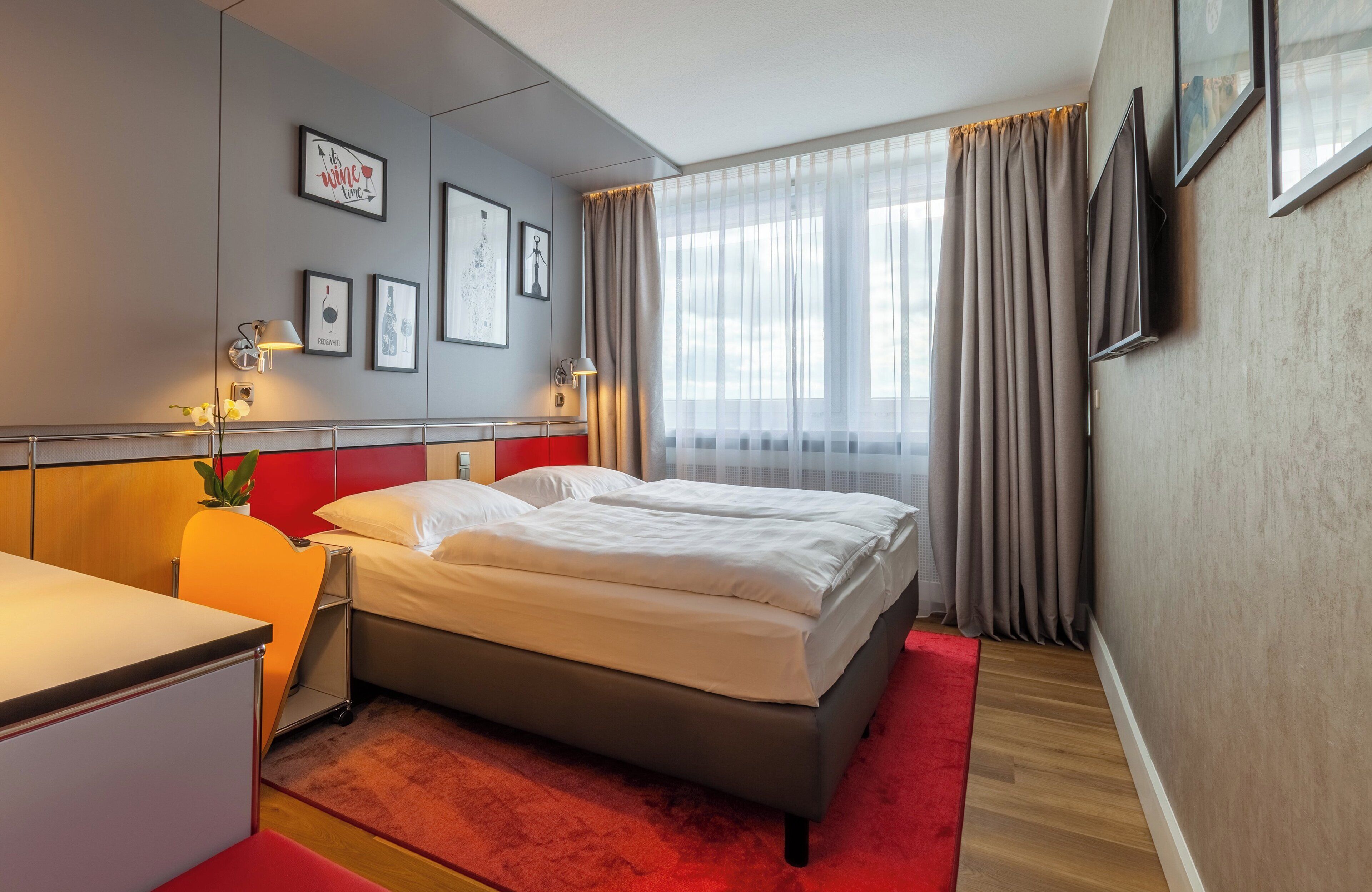 Une chambre d’hôtel avec un lit, un bureau, une chaise, une moquette rouge et une fenêtre avec des rideaux.