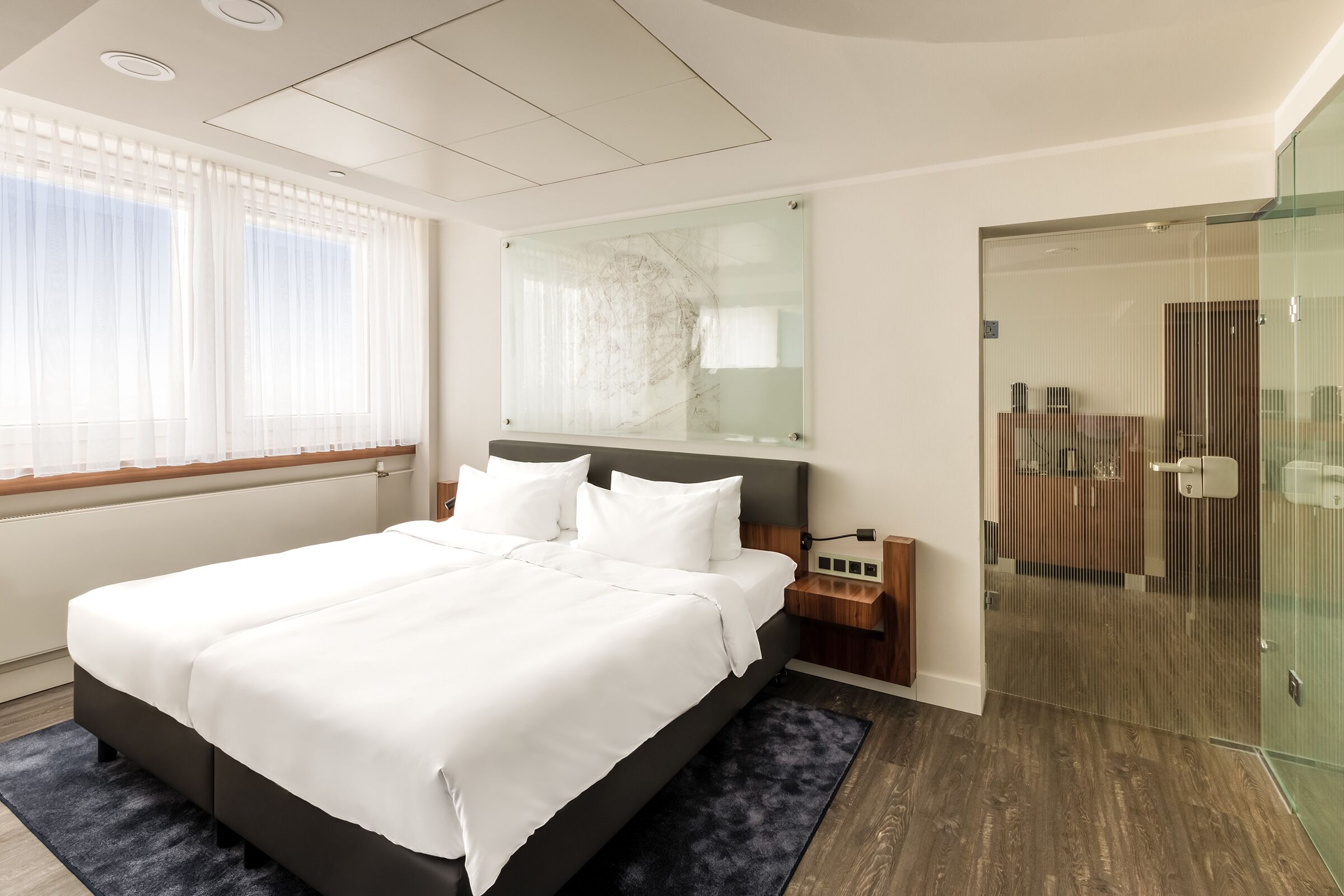 A modern hotel room with a large bed, a glass shower enclosure, and a view of the city.