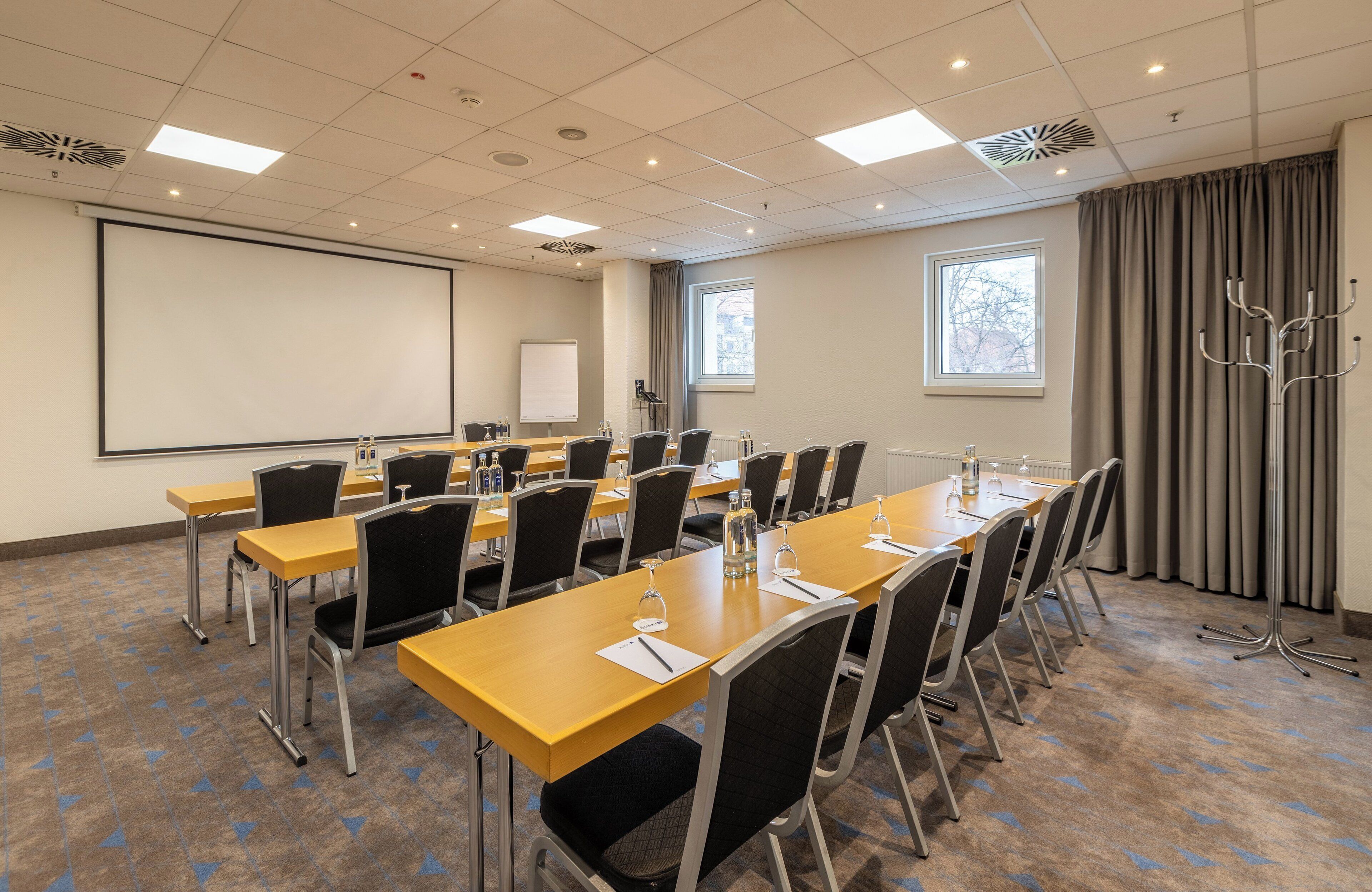 A conference room with a large screen, multiple tables, and chairs, equipped with water bottles and glasses.