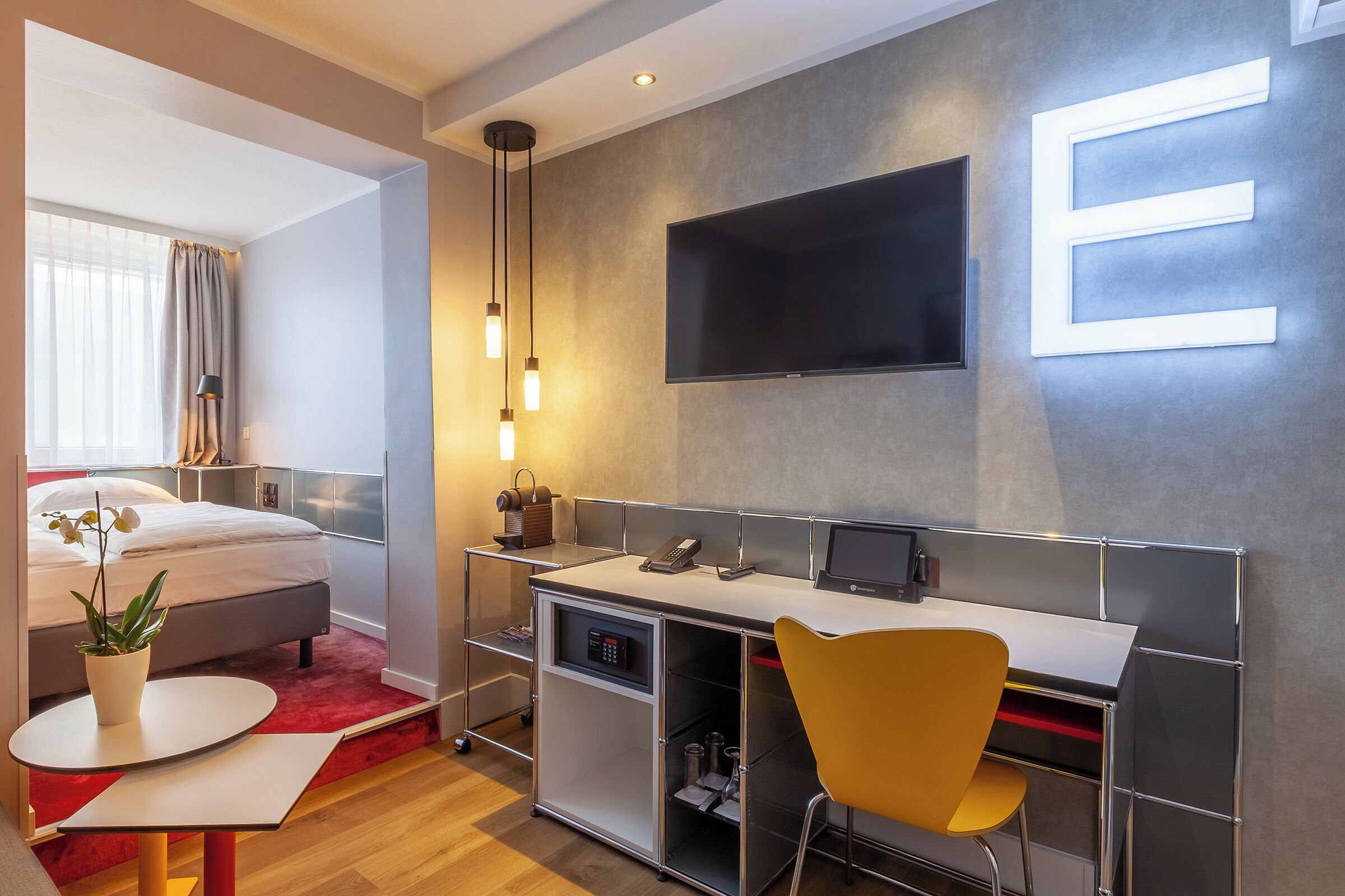 A modern hotel room with a bed, desk, chair, TV, and a potted plant.