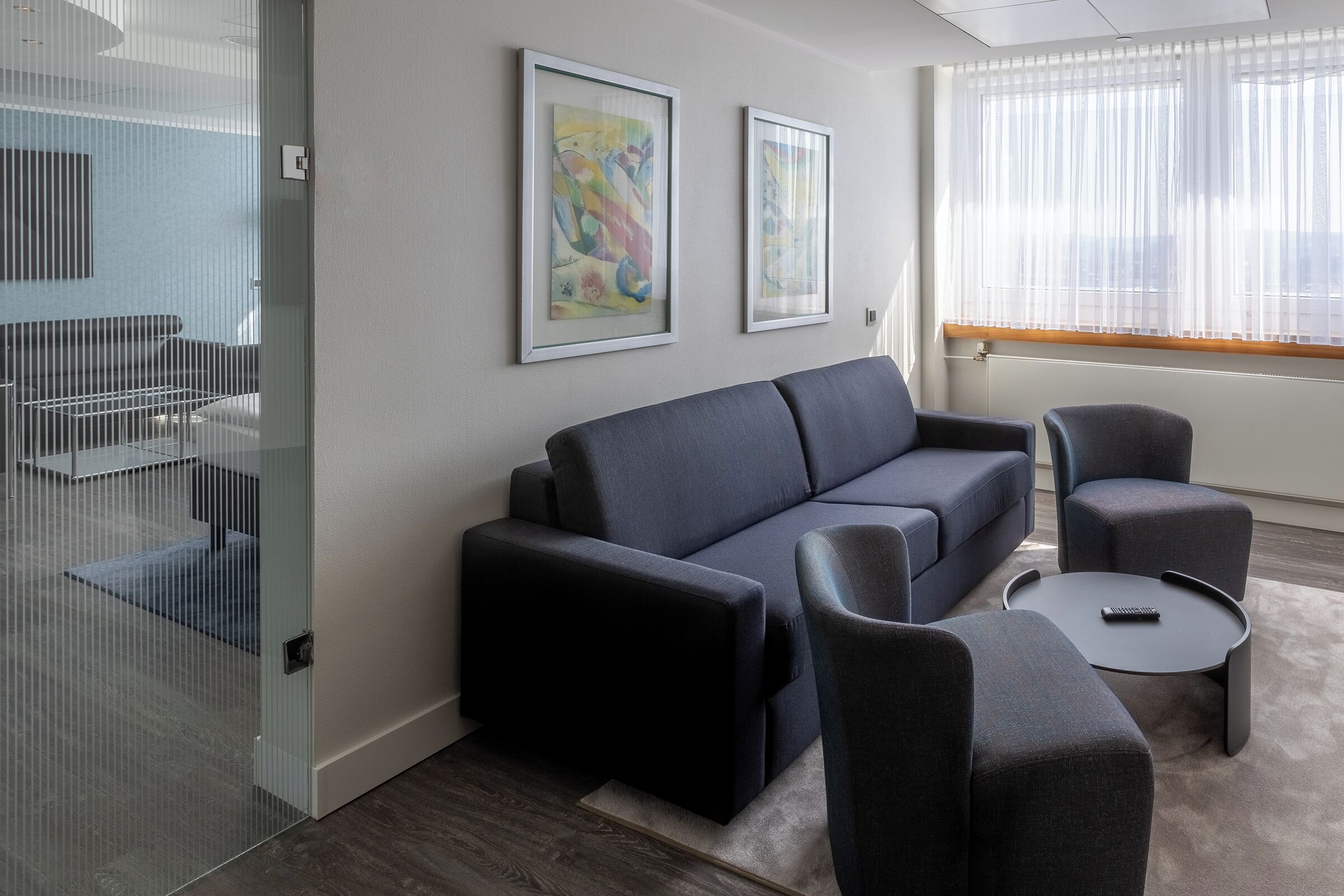 A modern living room with a dark sofa, two armchairs, and a small coffee table. There are two framed abstract paintings on the wall.