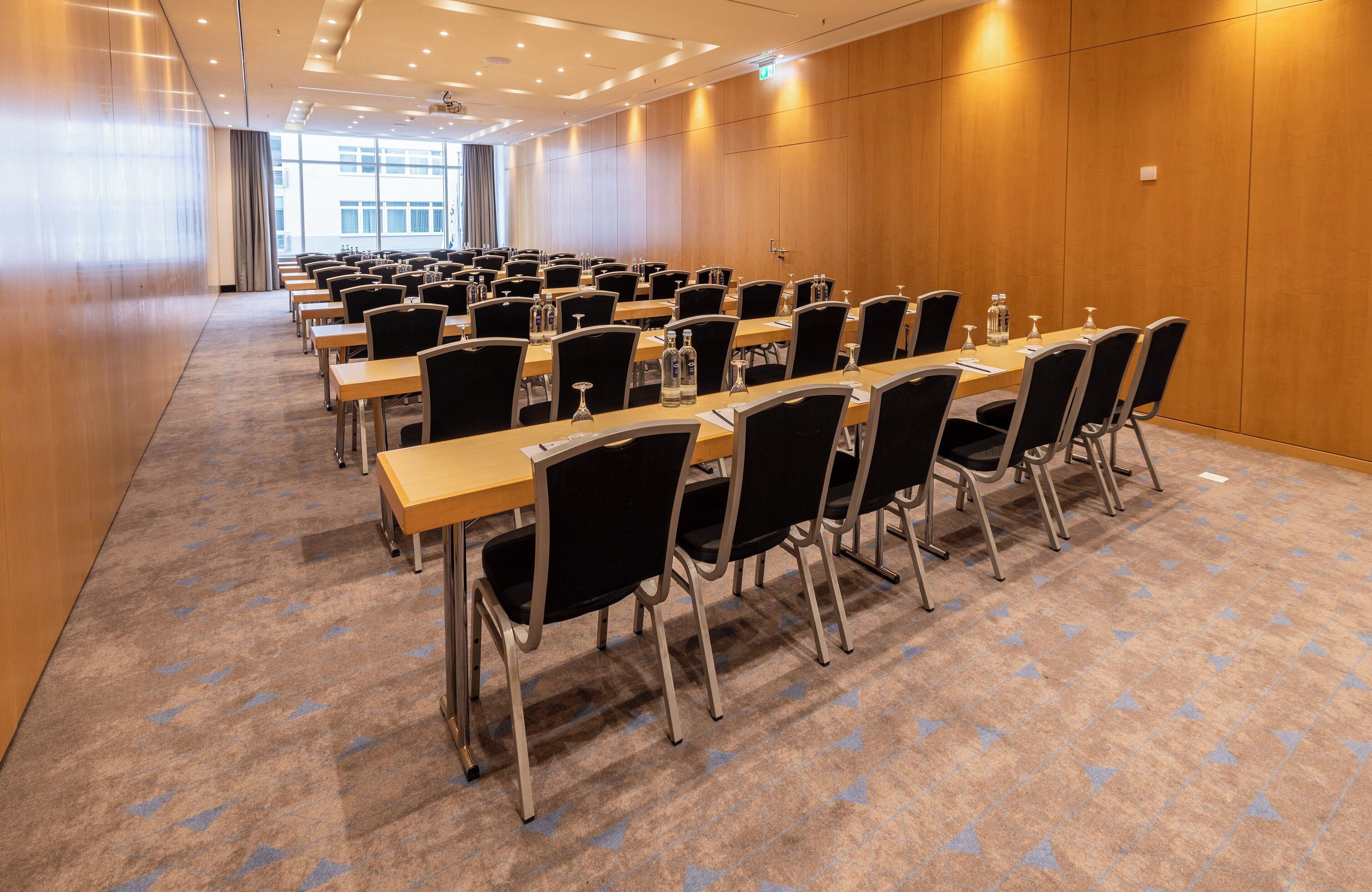 Un salón de conferencias con filas de mesas y sillas, paredes de paneles de madera y suelo alfombrado.