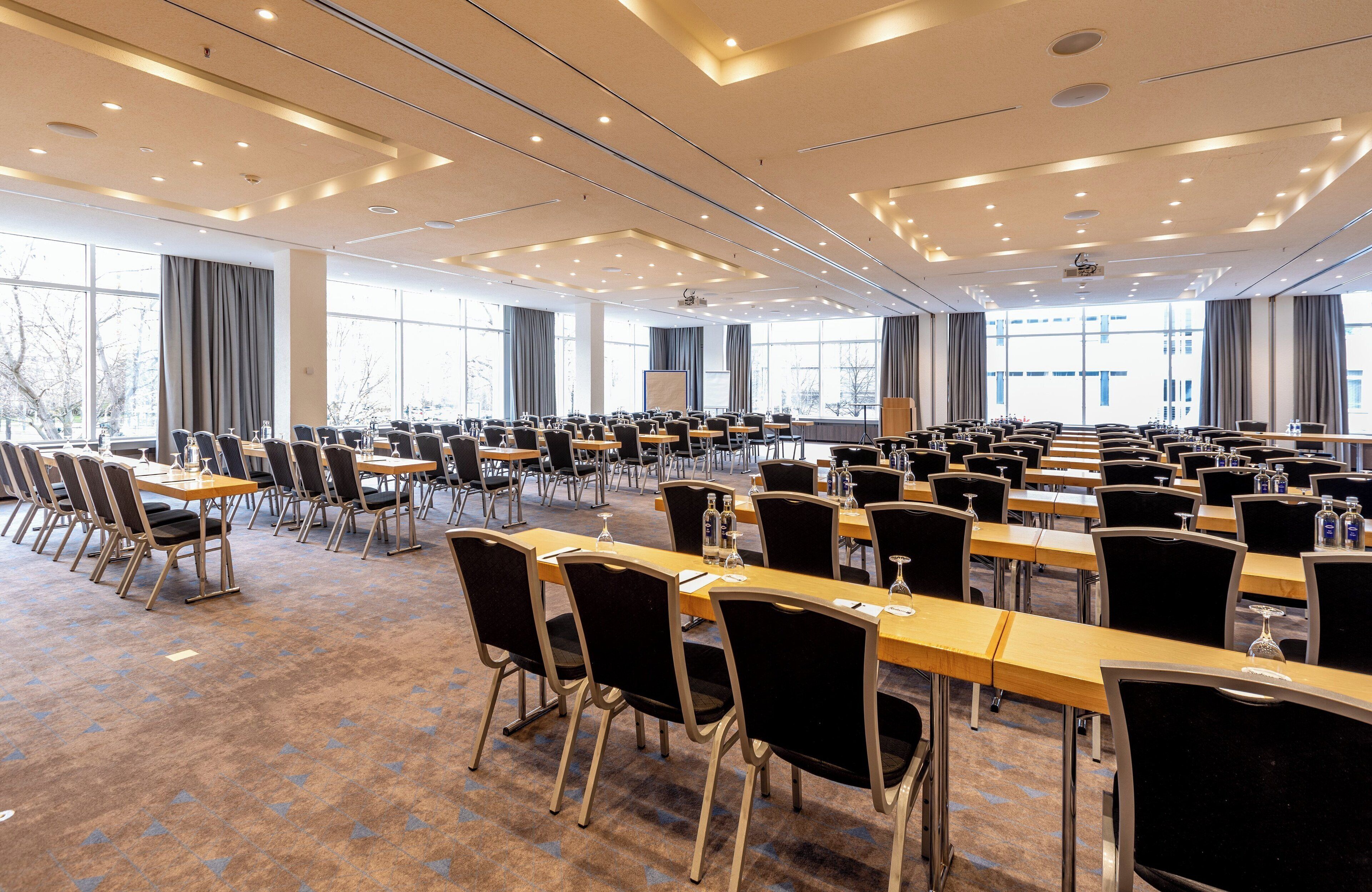 Ein großer Konferenzraum mit Tischen, Stühlen und einem großen Fensterfronten sowie einer Projektionsleinwand.