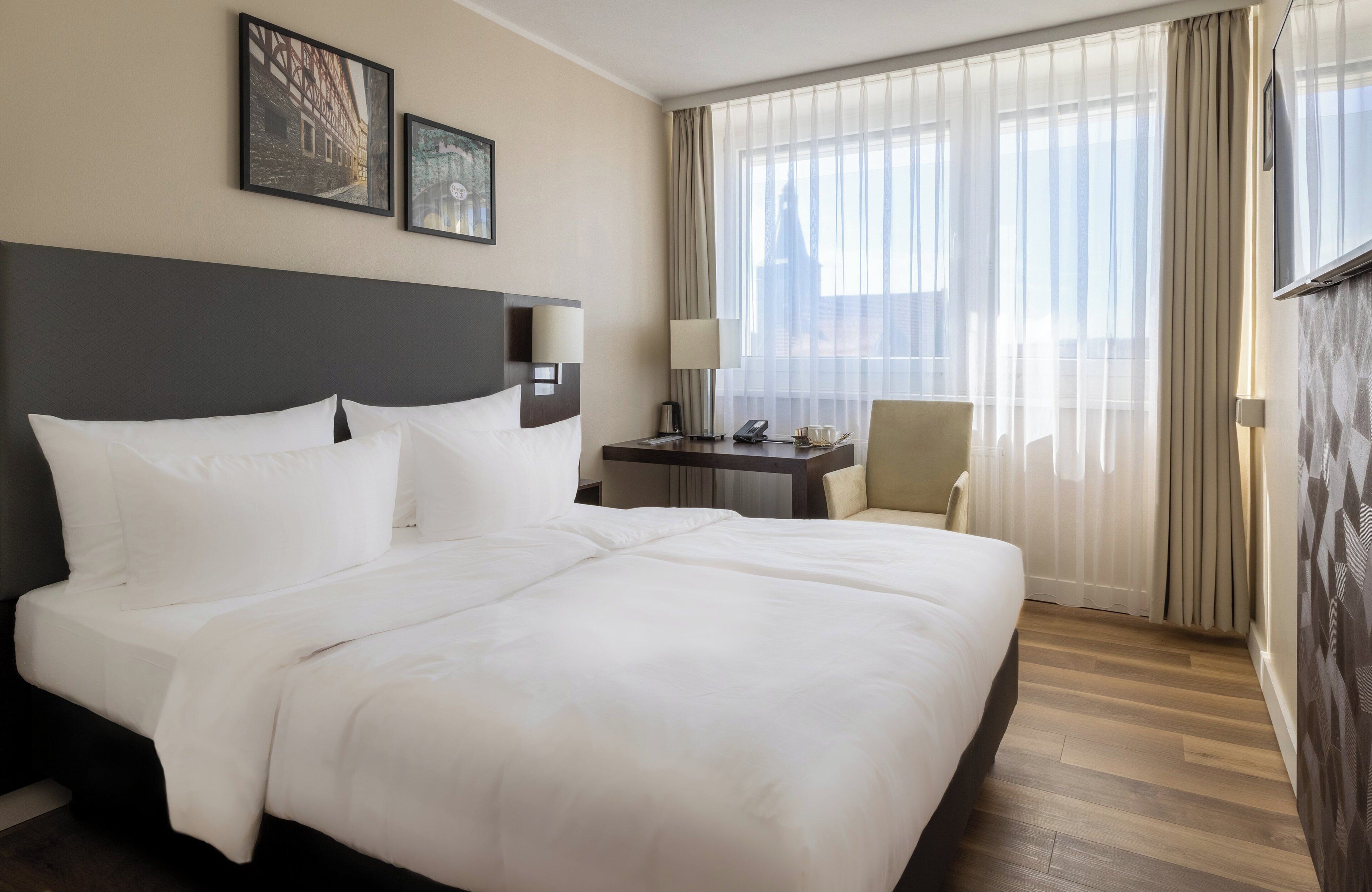A hotel room with a large bed, a desk with a lamp, a chair, and a window with curtains.