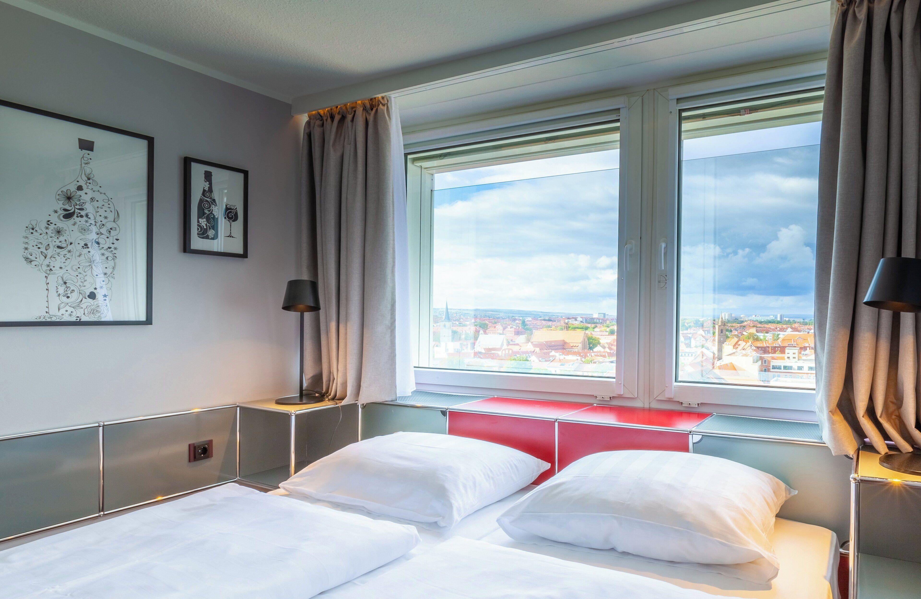 A hotel room with a bed, a nightstand, a lamp, and a window with a city view.