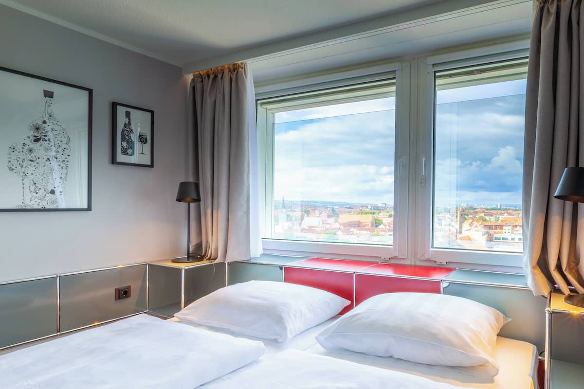A hotel room with a bed, a nightstand, a lamp, and a window with a city view.