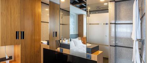 A modern hotel room with a wooden wardrobe, a large mirror, a bed, a bathroom with a glass shower, and a vanity with a sink and mirror.