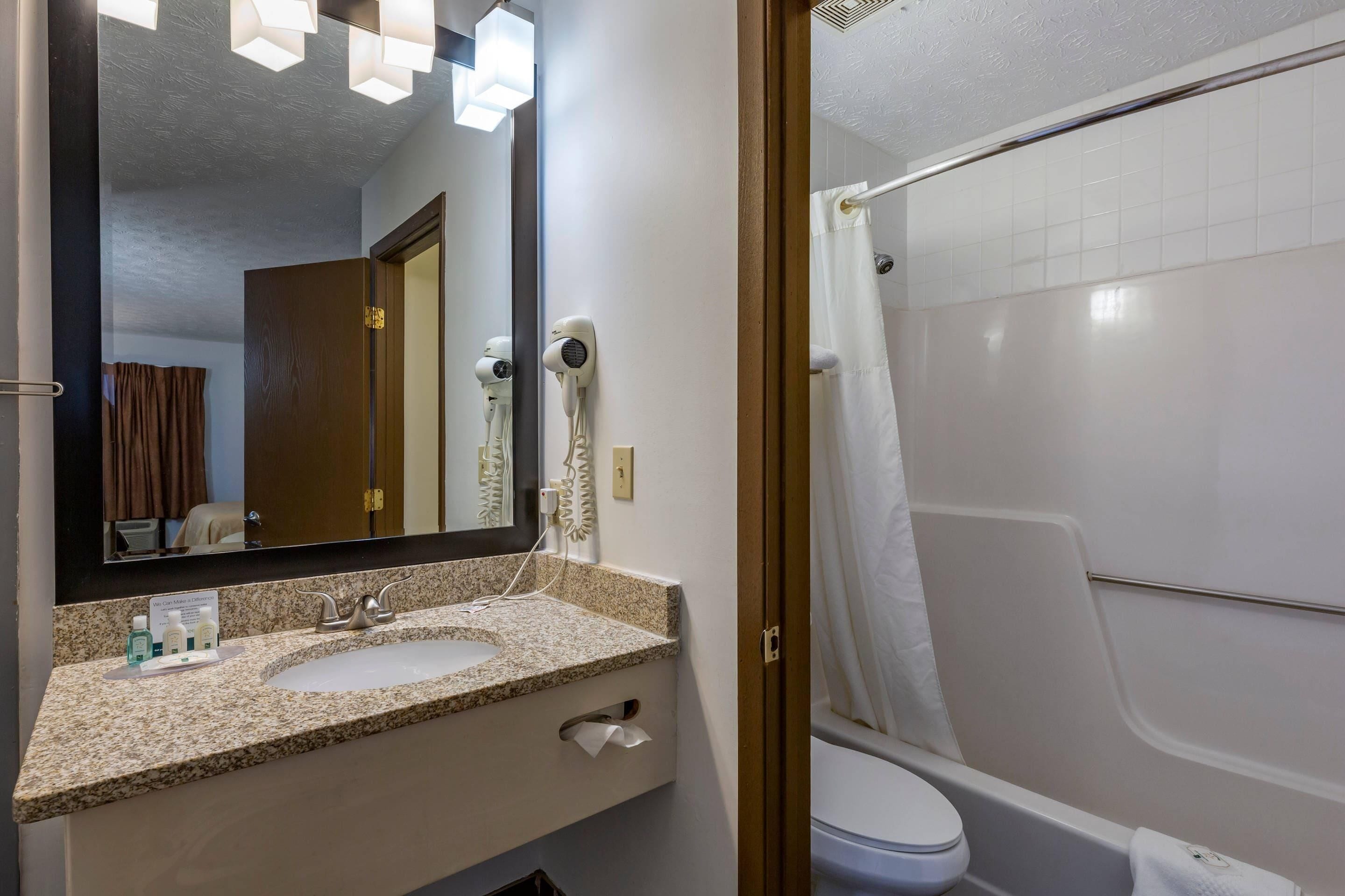 A bathroom with a granite countertop, a sink, a mirror, a hairdryer, and a shower area.