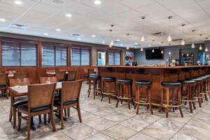 A restaurant interior with wooden tables and chairs, a bar area with stools, and a tiled floor.