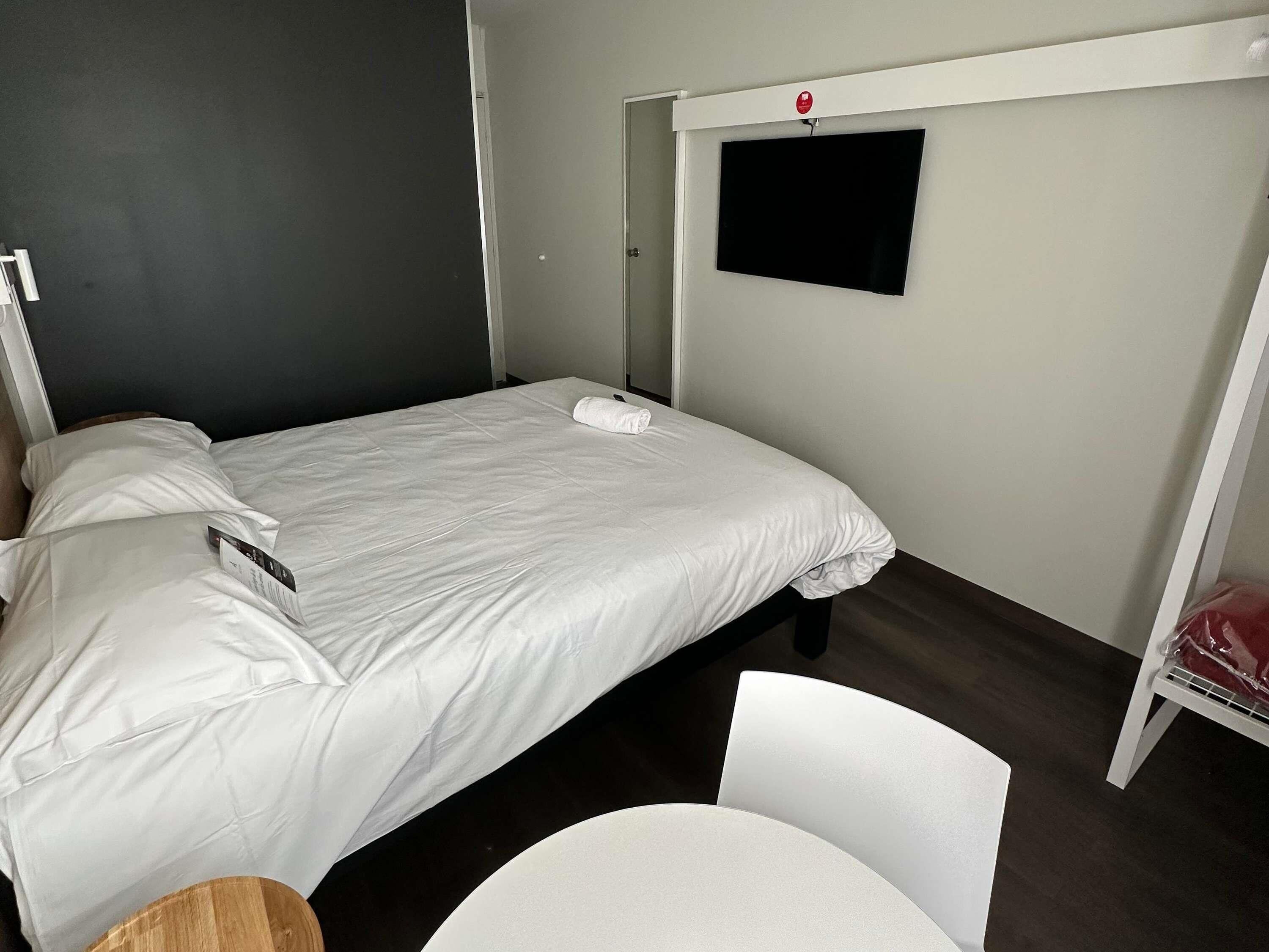 A single bed with white bedding, a small table, a chair, a TV mounted on the wall, and a door.