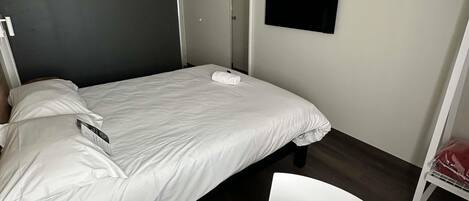 Una cama individual con ropa de cama blanca, una mesita pequeña, una silla, un televisor montado en la pared y una puerta.