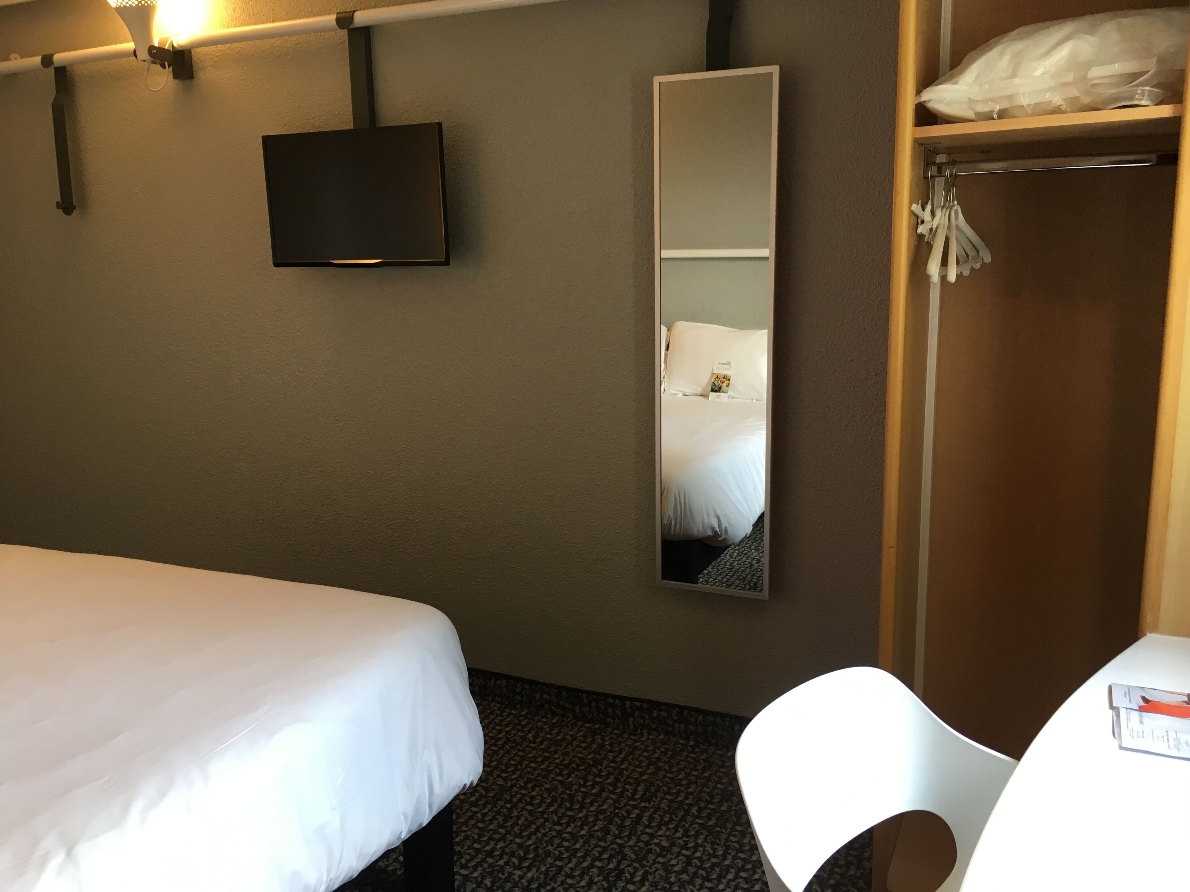 A hotel room with a single bed, a television mounted on the wall, a large mirror, a wardrobe, and a small table with a chair.