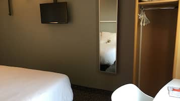 A hotel room with a single bed, a television mounted on the wall, a large mirror, a wardrobe, and a small table with a chair.