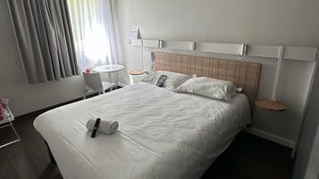 Quarto de hotel com uma cama grande, uma mesa de cabeceira, uma pequena mesa redonda, uma cadeira, uma janela com cortinas e um telefone fixado na parede.