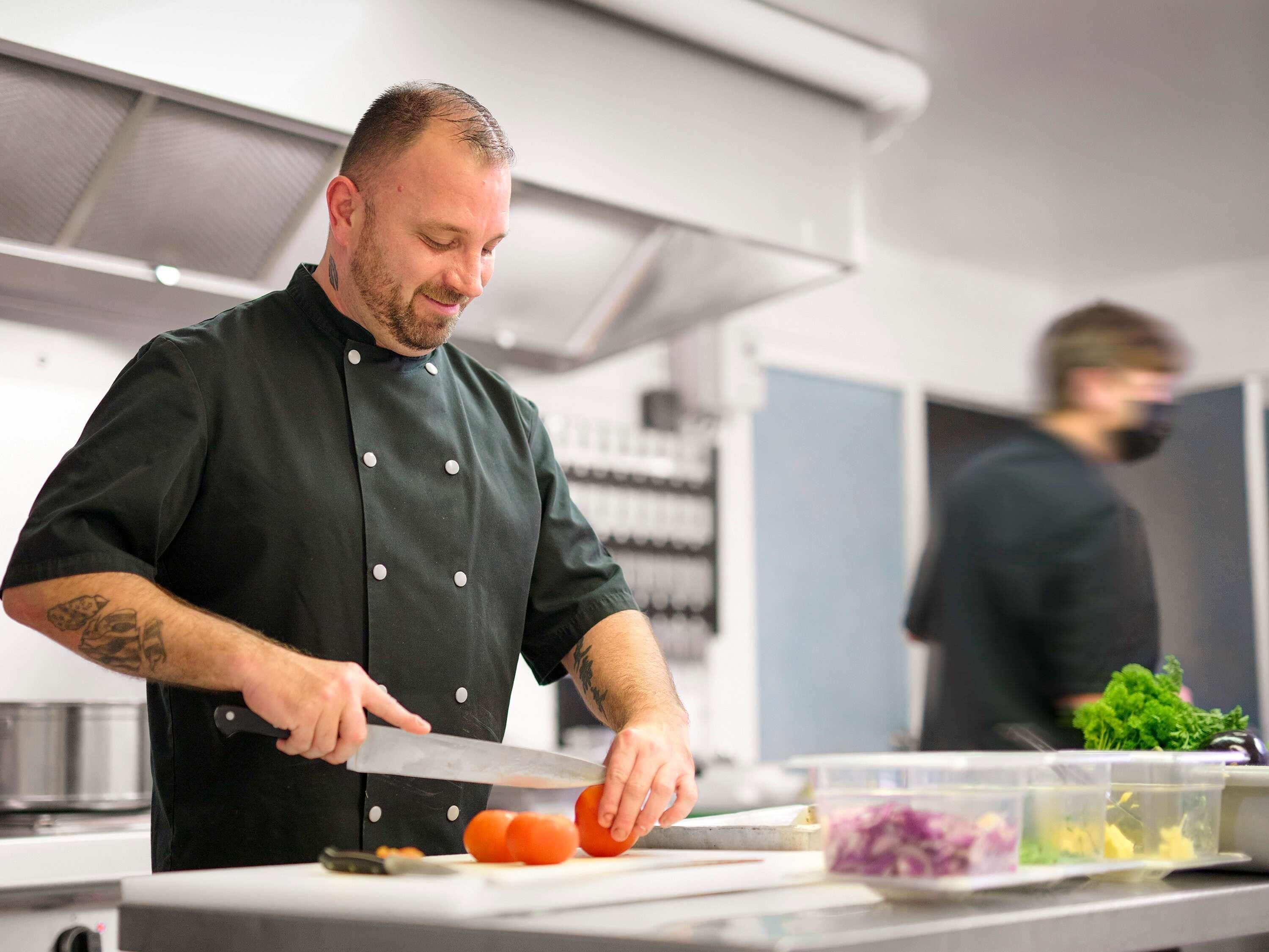 A chef in a black uniform with a tattoo on his arm, cutting a tomato on a white cutting board in a kitchen with stainless steel appliances.