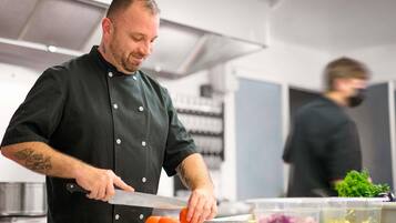 A chef in a black uniform with a tattoo on his arm, cutting a tomato on a white cutting board in a kitchen with stainless steel appliances.