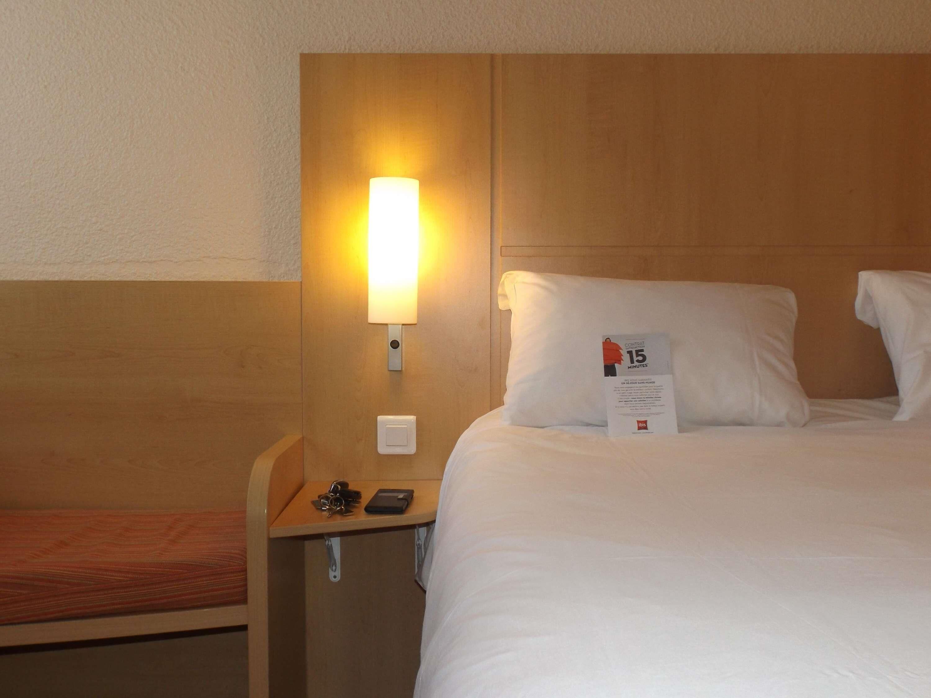 A hotel room with a wooden headboard, a bedside table with a lamp, a bed with white linens, and a pillow with a '15 minutes' sign.