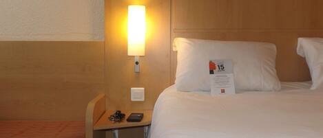 Habitación de hotel con cabecera de madera, una mesita de noche con lámpara, cama con ropa blanca y almohada con un cartel que dice '15 minutos'.