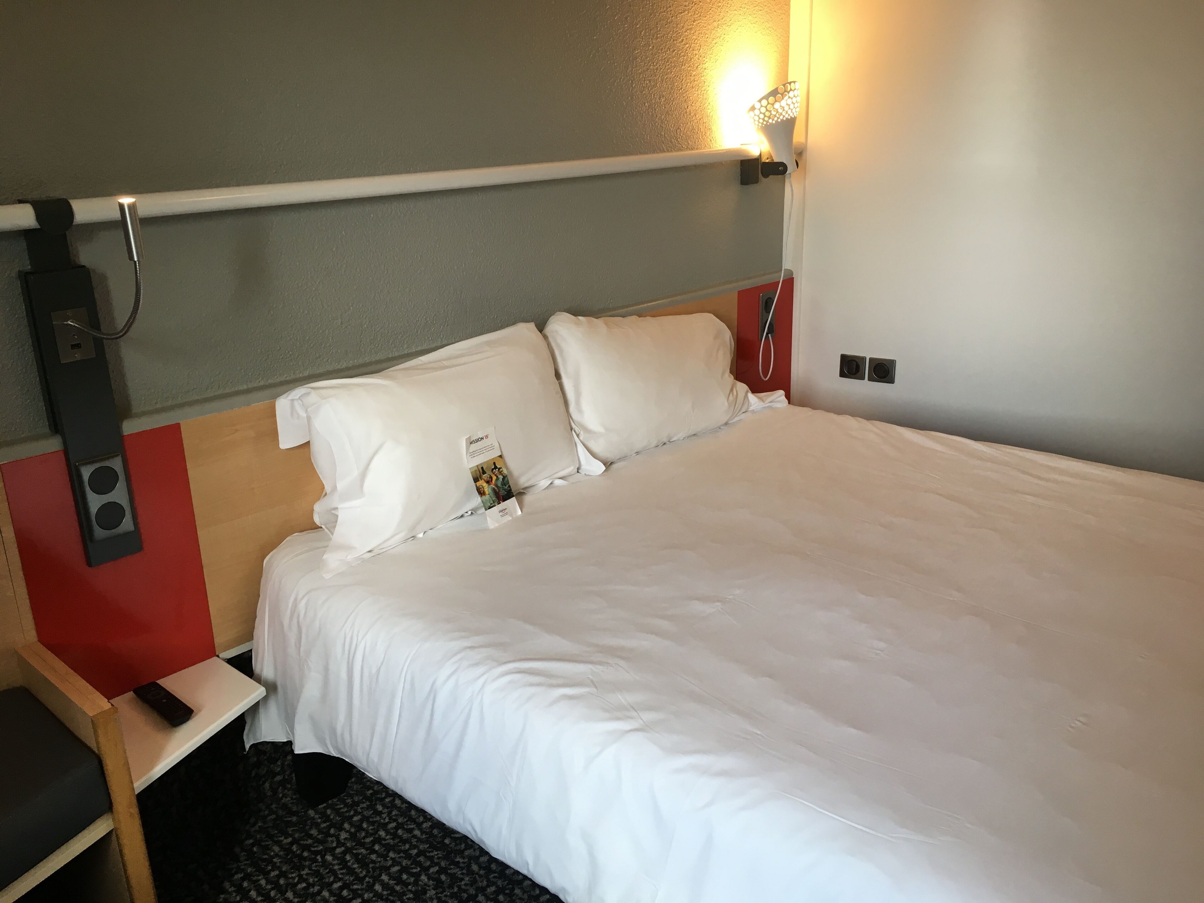 A hotel room with a bed, a bedside table, a lamp, and a wall-mounted light fixture.