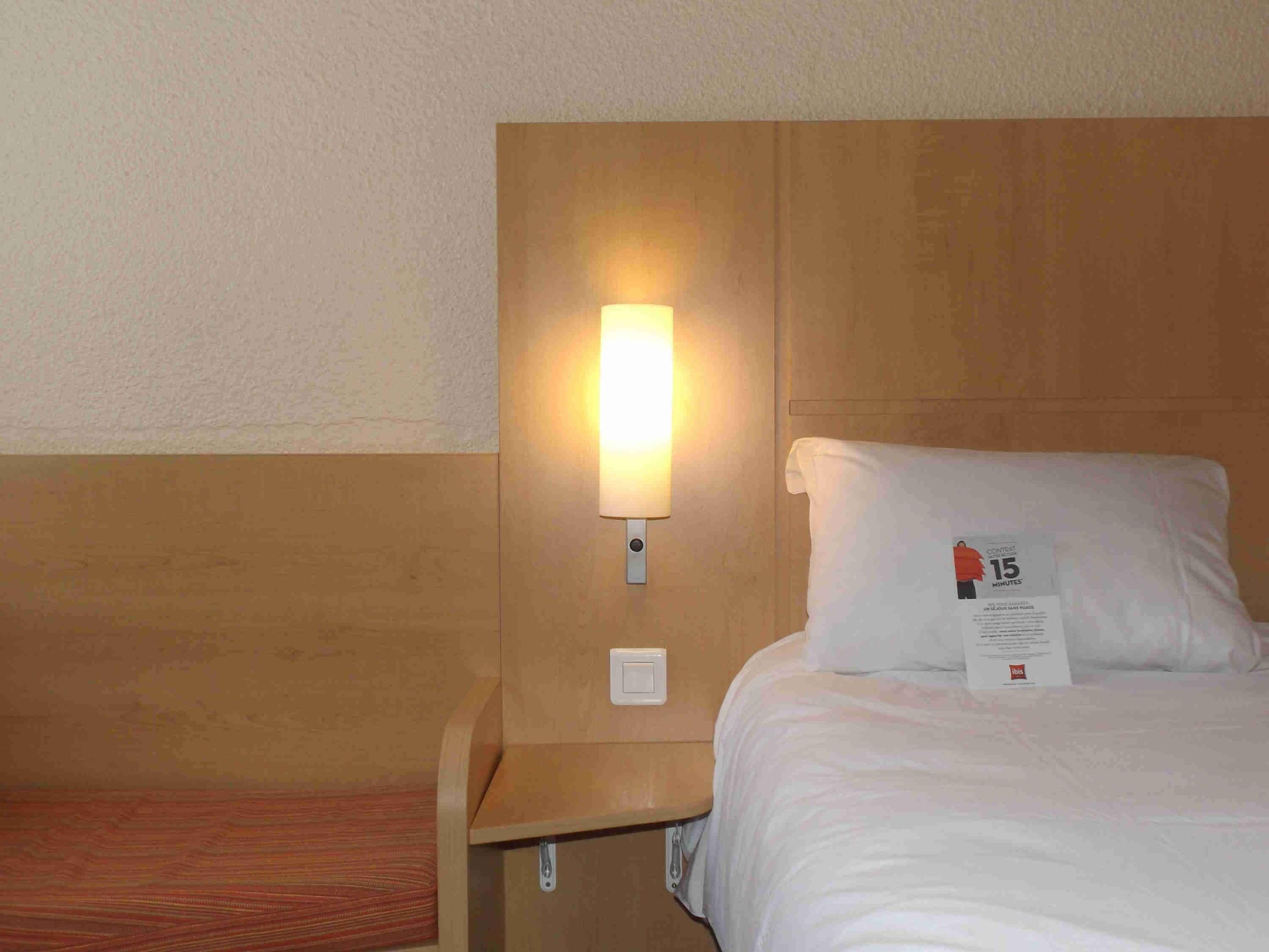A hotel room with a wooden headboard, a bedside lamp, a white pillow with a tag, and a small wooden nightstand.