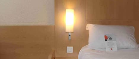 A hotel room with a wooden headboard, a bedside lamp, a white pillow with a tag, and a small wooden nightstand.