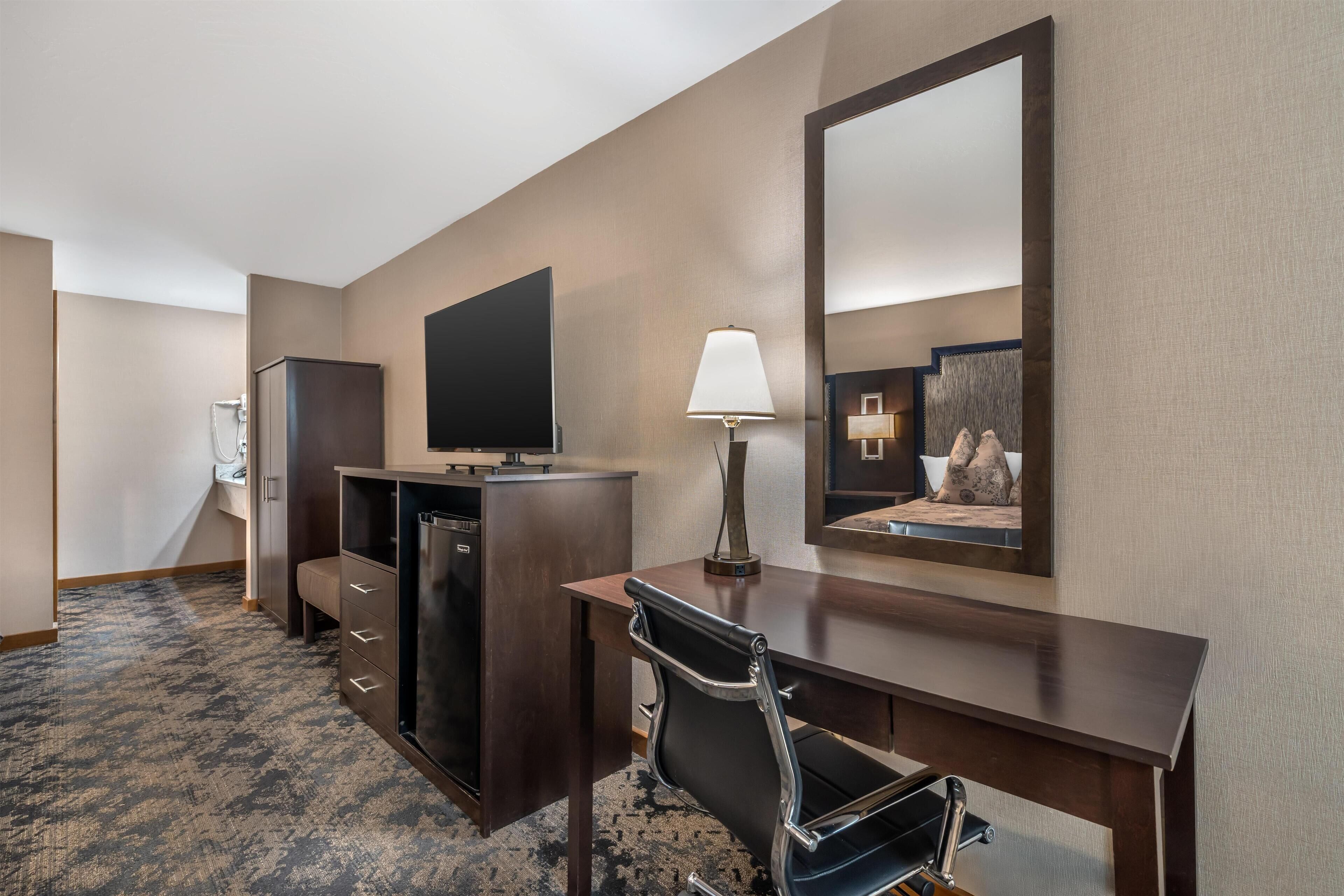 A hotel room with a desk, chair, television, and a mirror.