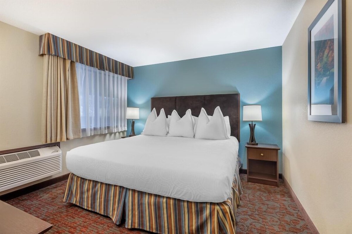 A hotel room with a bed, two bedside lamps, a window with curtains, a wall-mounted air conditioner, and a framed picture on the wall.
